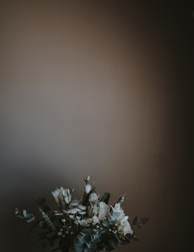 A bouquet of white flowers with green leaves is positioned against a muted, dark background. The arrangement appears delicate and elegant, with a focus on simplicity and natural elements.