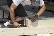 A person is kneeling on a construction site, assembling a wooden frame using clamps. The individual is wearing a cap, a white t-shirt, black shorts, and sneakers. The scene is outdoors, possibly in daylight, indicating work in progress.