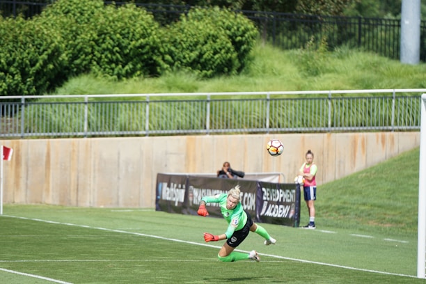 A soccer player in a green uniform dives to head a ball near the goal. There are several people watching in the background, including someone wearing a pink bib. The scene is set on a green grass field with a barrier and lush foliage visible in the background.