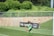 A soccer player in a green uniform dives to head a ball near the goal. There are several people watching in the background, including someone wearing a pink bib. The scene is set on a green grass field with a barrier and lush foliage visible in the background.