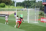 A soccer match is taking place with a player in a red jersey attempting a header near the goalpost. Other players in white jerseys and a goalkeeper in a green kit are nearby, focused on the ball in motion. In the background, there's green grass and a fence, with a photographer capturing the action.