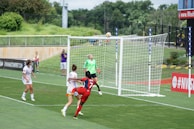A soccer match is taking place with a player in a red jersey attempting a header near the goalpost. Other players in white jerseys and a goalkeeper in a green kit are nearby, focused on the ball in motion. In the background, there's green grass and a fence, with a photographer capturing the action.