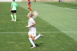 A female soccer player in a white and pastel-colored uniform is running on a grassy field. She has blonde hair tied up and is wearing white socks and cleats. In the background, another player in a green top and black shorts is visible, along with a player in a red and black uniform.