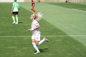 A female soccer player in a white and pastel-colored uniform is running on a grassy field. She has blonde hair tied up and is wearing white socks and cleats. In the background, another player in a green top and black shorts is visible, along with a player in a red and black uniform.
