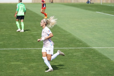 A female soccer player in a white and pastel-colored uniform is running on a grassy field. She has blonde hair tied up and is wearing white socks and cleats. In the background, another player in a green top and black shorts is visible, along with a player in a red and black uniform.