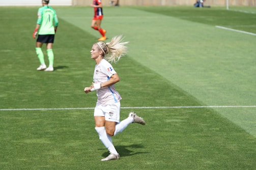 A female soccer player in a white and pastel-colored uniform is running on a grassy field. She has blonde hair tied up and is wearing white socks and cleats. In the background, another player in a green top and black shorts is visible, along with a player in a red and black uniform.
