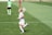 A female soccer player in a white and pastel-colored uniform is running on a grassy field. She has blonde hair tied up and is wearing white socks and cleats. In the background, another player in a green top and black shorts is visible, along with a player in a red and black uniform.