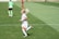 A female soccer player in a white and pastel-colored uniform is running on a grassy field. She has blonde hair tied up and is wearing white socks and cleats. In the background, another player in a green top and black shorts is visible, along with a player in a red and black uniform.