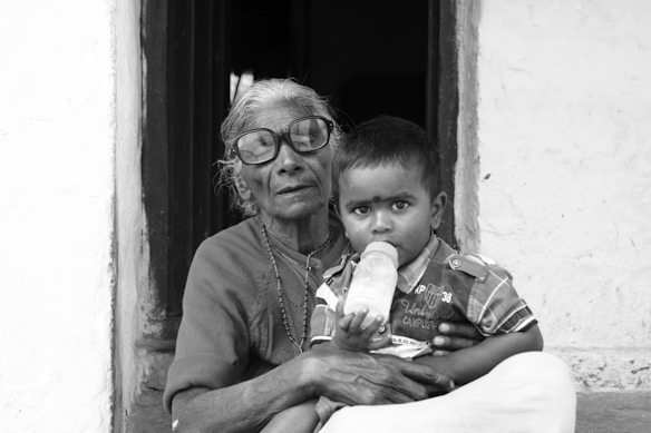 An elderly woman with glasses and a traditional necklace holds a young child sitting on her lap. The child is drinking from a bottle and wearing a striped shirt. They are seated in the doorway of a rustic setting.