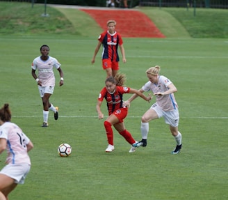 A group of female soccer players are engaged in an intense match on a green field. One player in a red and black uniform is in possession of the ball while trying to evade an opponent wearing a white and blue uniform. Other players are seen in the background, actively participating and focusing on the ongoing play.
