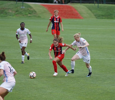 A group of female soccer players are engaged in an intense match on a green field. One player in a red and black uniform is in possession of the ball while trying to evade an opponent wearing a white and blue uniform. Other players are seen in the background, actively participating and focusing on the ongoing play.
