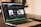 A laptop displaying a music streaming application on its screen. The application shows various playlists such as Focus, Deep Focus, Chill and Study Beats, and Productive Morning. Next to the laptop, there is a black notebook titled 'The ONE Thing' and a smartphone placed on top of it. The background is slightly blurred, suggesting an indoor setting with warm lighting.
