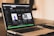 A laptop displaying a music streaming application on its screen. The application shows various playlists such as Focus, Deep Focus, Chill and Study Beats, and Productive Morning. Next to the laptop, there is a black notebook titled 'The ONE Thing' and a smartphone placed on top of it. The background is slightly blurred, suggesting an indoor setting with warm lighting.