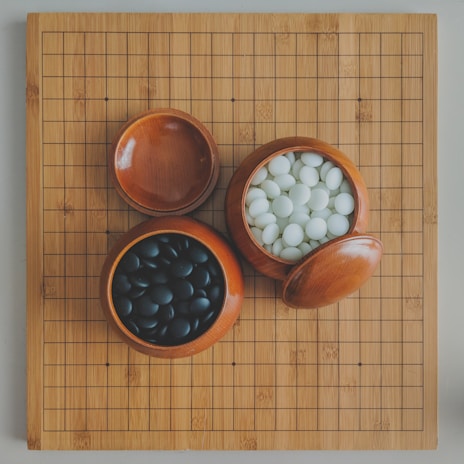 A wooden board with a grid pattern accompanies two open wooden bowls; one containing black stones and the other white stones, typically used for playing the game Go.