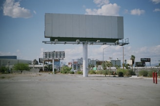 An empty billboard stands prominently in a sparse urban environment, surrounded by a few scattered palm trees and patches of dry grass. The sky is partly cloudy, providing a bright backdrop, while other billboards and buildings are visible in the distance.