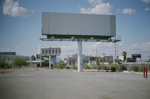 An empty billboard stands prominently in a sparse urban environment, surrounded by a few scattered palm trees and patches of dry grass. The sky is partly cloudy, providing a bright backdrop, while other billboards and buildings are visible in the distance.