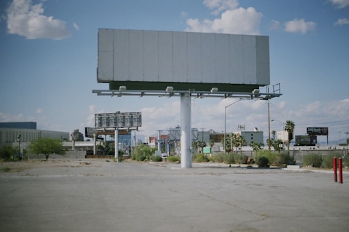 An empty billboard stands prominently in a sparse urban environment, surrounded by a few scattered palm trees and patches of dry grass. The sky is partly cloudy, providing a bright backdrop, while other billboards and buildings are visible in the distance.