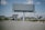 An empty billboard stands prominently in a sparse urban environment, surrounded by a few scattered palm trees and patches of dry grass. The sky is partly cloudy, providing a bright backdrop, while other billboards and buildings are visible in the distance.