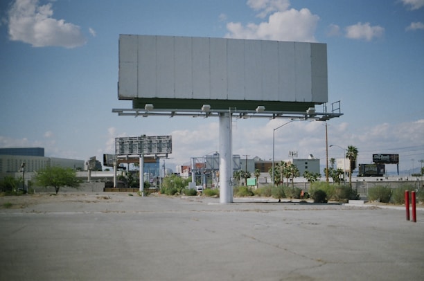 An empty billboard stands prominently in a sparse urban environment, surrounded by a few scattered palm trees and patches of dry grass. The sky is partly cloudy, providing a bright backdrop, while other billboards and buildings are visible in the distance.