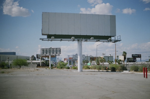 An empty billboard stands prominently in a sparse urban environment, surrounded by a few scattered palm trees and patches of dry grass. The sky is partly cloudy, providing a bright backdrop, while other billboards and buildings are visible in the distance.
