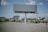 An empty billboard stands prominently in a sparse urban environment, surrounded by a few scattered palm trees and patches of dry grass. The sky is partly cloudy, providing a bright backdrop, while other billboards and buildings are visible in the distance.