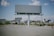 An empty billboard stands prominently in a sparse urban environment, surrounded by a few scattered palm trees and patches of dry grass. The sky is partly cloudy, providing a bright backdrop, while other billboards and buildings are visible in the distance.