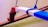 A close-up of a boat's outrigger with a wooden beam painted in red, white, and blue colors. The beam is secured with bright blue ropes, and the background shows calm, greenish water.