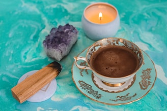 A delicate porcelain teacup filled with dark coffee sits on a saucer decorated with intricate golden designs. Nearby, a lit candle in a simple holder casts a warm glow, complementing a piece of raw amethyst crystal. A small wooden incense stick rests on a circular base, contributing to the tranquil setting. The background is a swirling pattern of turquoise hues.