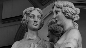 A black and white image of a classical sculpture featuring three figures. The focus is on two prominently displayed busts, both exhibiting intricate hairstyles and serene expressions. The background includes architectural elements with an archway.