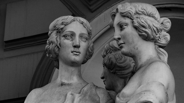 A black and white image of a classical sculpture featuring three figures. The focus is on two prominently displayed busts, both exhibiting intricate hairstyles and serene expressions. The background includes architectural elements with an archway.