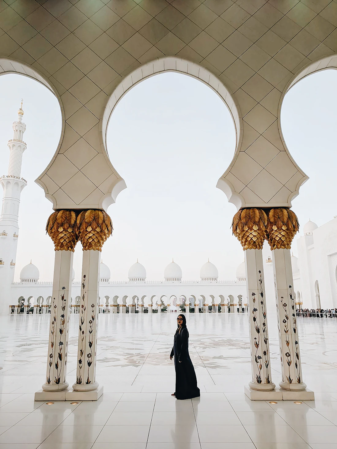 A model in a black abaya beneath the arches of a grand white mosque.