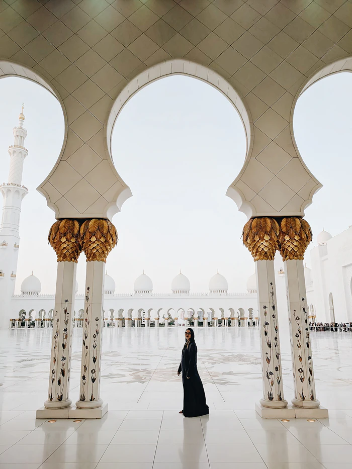 A model in a black abaya beneath the arches of a grand white mosque.