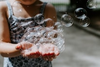 Close-up of a child’s hands holding a bright yellow bubble wand with bubbles floating around.