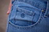 A close-up view of the back pocket of a pair of blue denim jeans. The small pocket displays a subtle emblem embossed on the fabric. Visible stitching and a metal rivet highlight the classic jean design.