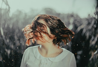A person with short hair, adorned with specks of golden glitter, is captured in mid-motion as their hair swirls around their face. The background consists of a blurred outdoor setting, suggesting a natural environment.