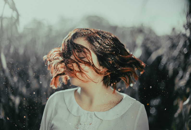 A person with short hair, adorned with specks of golden glitter, is captured in mid-motion as their hair swirls around their face. The background consists of a blurred outdoor setting, suggesting a natural environment.