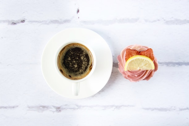 A cup of dark coffee is placed in a white saucer on a light surface next to a cupcake. The cupcake is topped with pink frosting, a slice of lemon, and a small decorative piece, possibly bacon.