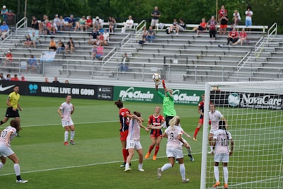 A competitive women's soccer match is taking place, with players actively engaged around the goal area. The goalkeeper in a green jersey is jumping to catch the ball, while players wearing white and red jerseys closely contend for position. The background shows a partially filled spectator stand, with fans watching the game.