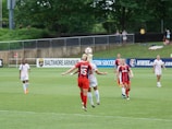 A soccer match is taking place on a grassy field with several female players actively engaged in the game. Two players are in the foreground, with one wearing a red and navy striped jersey jumping to head the ball, while another player, in a white uniform, is contesting for the ball. Other players in various action poses are seen in the background, with a referee standing by the sidelines. Advertisements and banners are placed near the field, and a few spectators watch the game from behind a fence.