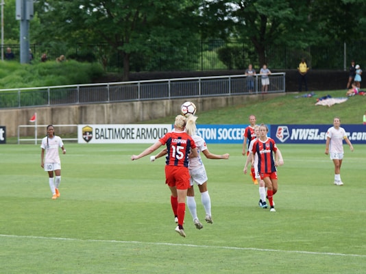 A soccer match is taking place on a grassy field with several female players actively engaged in the game. Two players are in the foreground, with one wearing a red and navy striped jersey jumping to head the ball, while another player, in a white uniform, is contesting for the ball. Other players in various action poses are seen in the background, with a referee standing by the sidelines. Advertisements and banners are placed near the field, and a few spectators watch the game from behind a fence.