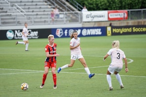 A soccer match with three female players in action on the field. One player in a red and navy striped uniform is moving towards the ball, while two players in white uniforms are defending. The scene is set in a stadium with empty stands and advertisements visible in the background.