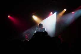 A person stands on stage in front of a microphone, surrounded by a dimly lit audience. Bright colored lights, including red, yellow, and white, create dramatic beams that illuminate the scene from above.