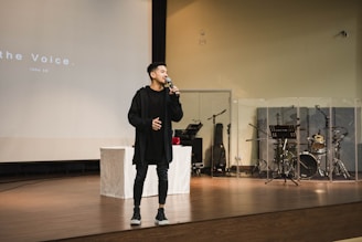 A person stands on a stage holding a microphone, appearing to speak or sing. Behind them, musical instruments like drums and music stands can be seen, partially enclosed by transparent panels. The stage is set in a large room with a wooden floor and a projection screen displaying text in the background.
