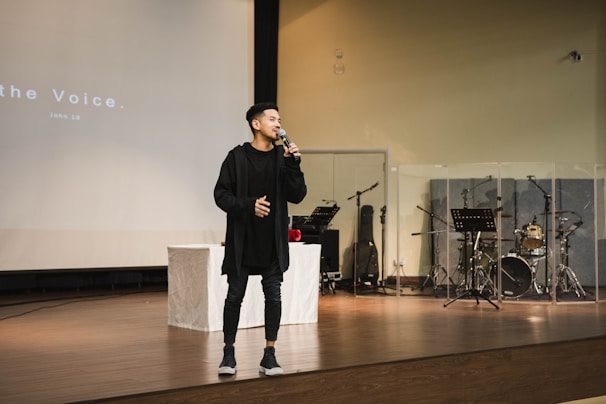 A person stands on a stage holding a microphone, appearing to speak or sing. Behind them, musical instruments like drums and music stands can be seen, partially enclosed by transparent panels. The stage is set in a large room with a wooden floor and a projection screen displaying text in the background.