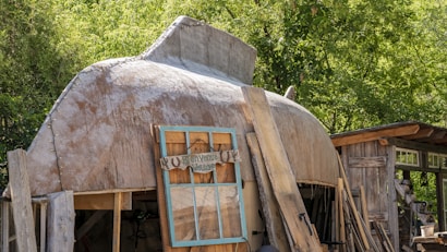 A rustic structure with a curved, dome-like roof and wooden panels leaning against it. There's an old window frame with a sign attached, surrounded by lush green foliage in the background. The setting appears to be outdoors, with various wooden elements giving a handcrafted, natural feel.