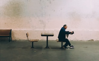 A solitary man sits on a bench in a spacious, minimally decorated room with aged walls. He appears relaxed and is focused on a portable device in his hands. The environment seems sparse, with only a few benches and chairs visible.