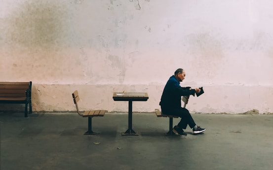 A solitary man sits on a bench in a spacious, minimally decorated room with aged walls. He appears relaxed and is focused on a portable device in his hands. The environment seems sparse, with only a few benches and chairs visible.