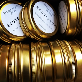 A collection of gold-colored metallic tins arranged closely together, some displaying labels that read '28 Days Program' and 'ECOTEATOX'. The tins have a shiny surface, highlighting their clean and polished appearance.
