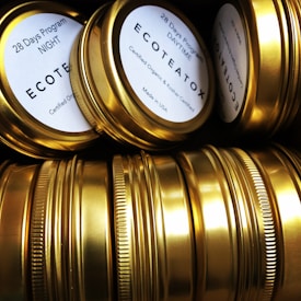 A collection of gold-colored metallic tins arranged closely together, some displaying labels that read '28 Days Program' and 'ECOTEATOX'. The tins have a shiny surface, highlighting their clean and polished appearance.