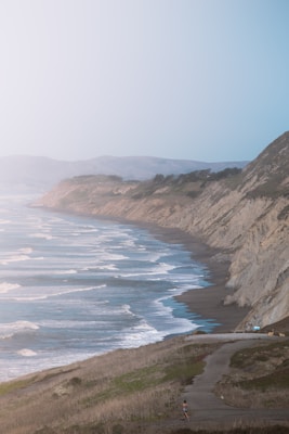A scenic coastal view features steep cliffs along a shoreline with waves gently crashing onto the sand. The landscape is bathed in soft light, enhancing the tranquil atmosphere. A lone person walks on a path that winds along the edge of the coast, adding a sense of scale and solitude to the expansive natural surroundings.